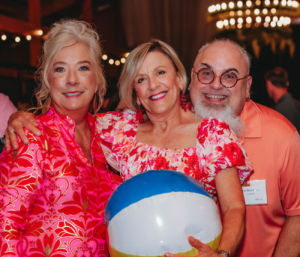 Three smiling adults pose together at a festive event, wearing vibrant pink floral shirts; a blue-and-white beach ball is held in front of them.