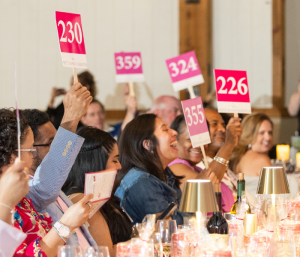 Guests raising pink bidding paddles with numbers, smiling and chatting.